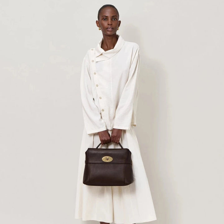 En kvinde i en hvid kjole holder en mørkebrun læderhåndtaske med guldspænde Model SKU-nummer
