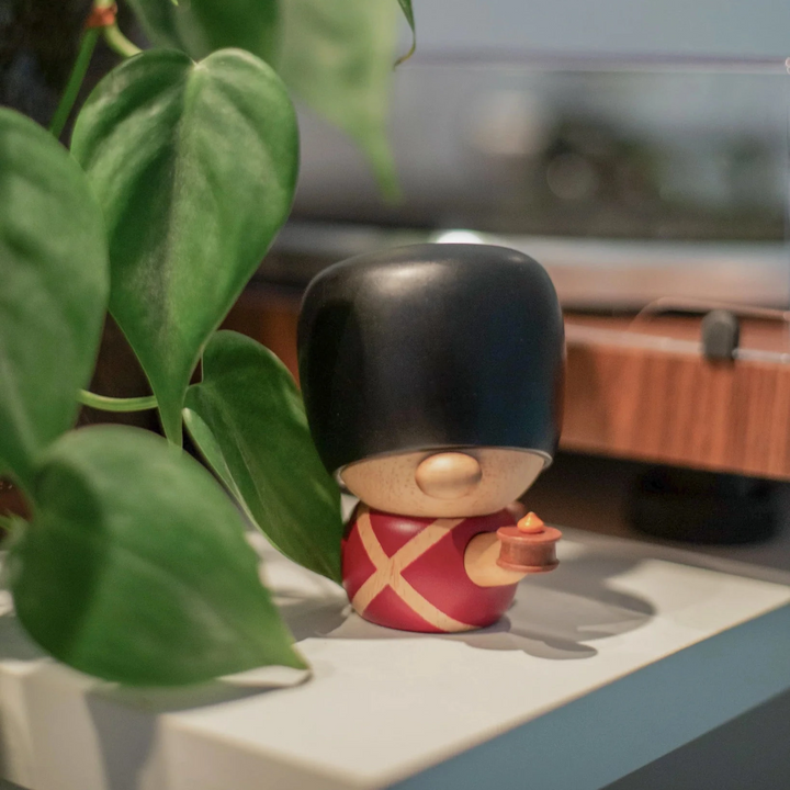 Fablewood HC Andersen's Fyrtøjet-8005-1 træfigur af garder i rød uniform og sort bjørneskinshue på hylde ved grøn plante. Model 8005-1