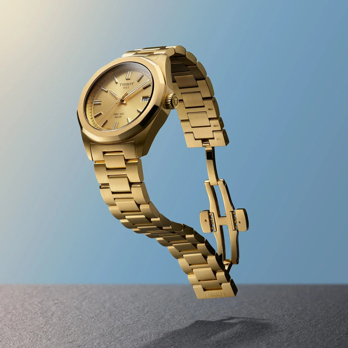 Tissot PRC 100 Solar 34mm guld PVD ur med gylden urskive og stållænke vist mod blå baggrund. Model T1518223302100