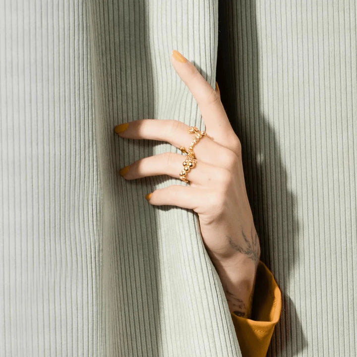 Vincent Orb Inferior Forgyldt Ring, vist på hånd med gul neglelak foran lysegrøn fløjlsgardin. Model VJ215-URG-001-2